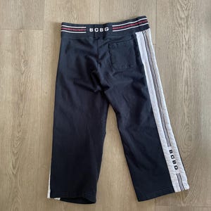 May include: Black sweatpants featuring a wide waistband with red and white stripes and the text BCBG. The pants include a single back pocket and white side panels with vertical stripes and the text BCBG printed near the bottom hem.