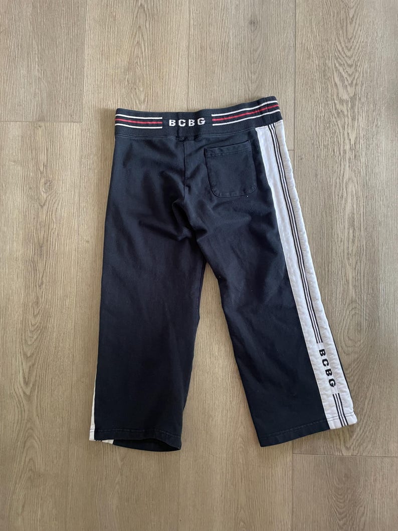 May include: Black sweatpants featuring a wide waistband with red and white stripes and the text BCBG. The pants include a single back pocket and white side panels with vertical stripes and the text BCBG printed near the bottom hem.