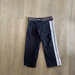 May include: Navy blue sweatpants featuring a white side stripe and the text BCBG printed on the waistband and lower leg. The pants include a back pocket and a waistband with red and white horizontal stripes. The design is simple and functional.