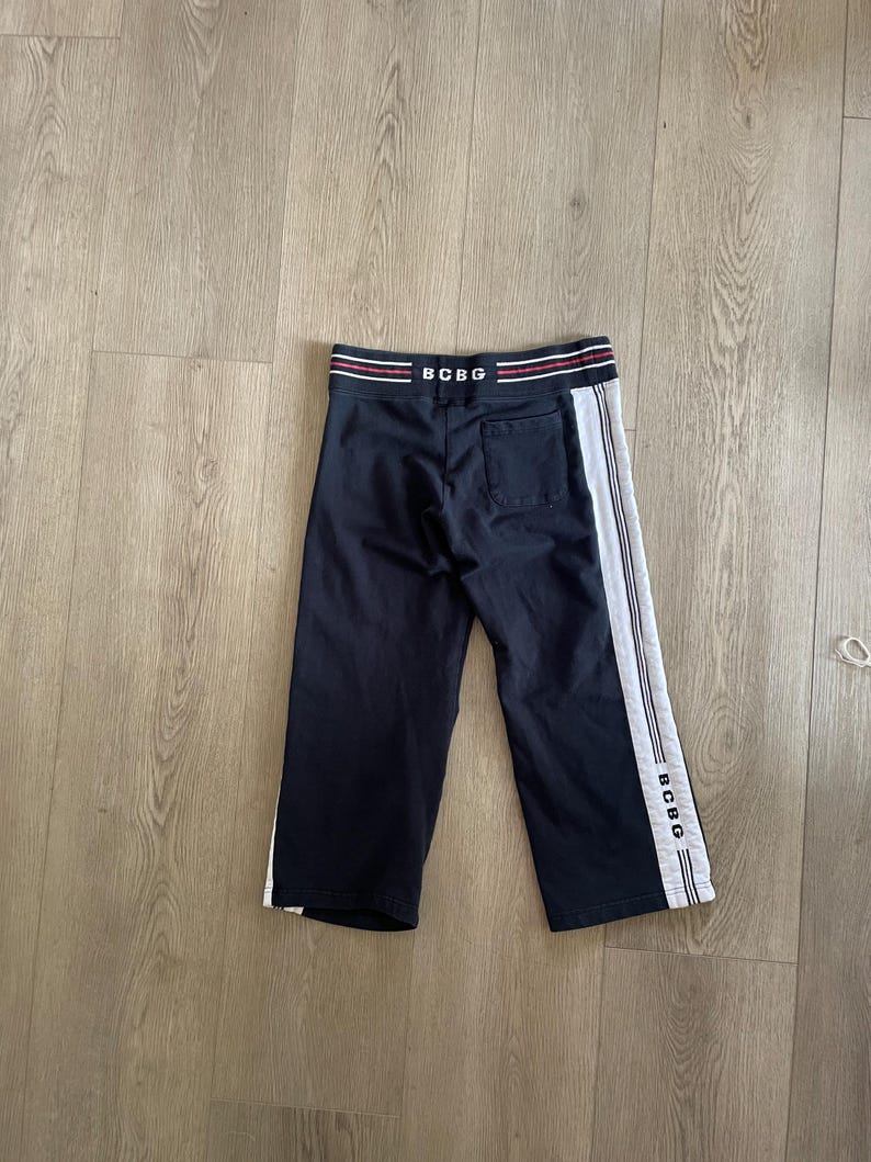 May include: Navy blue sweatpants featuring a white side stripe and the text BCBG printed on the waistband and lower leg. The pants include a back pocket and a waistband with red and white horizontal stripes. The design is simple and functional.