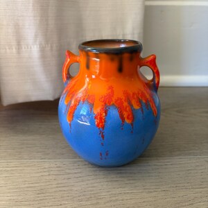 May include: A small, ceramic vase with a rounded body and two handles. The vase is painted with a vibrant color scheme: blue at the base, transitioning to orange, and a dark brown rim. The orange paint has red accents.