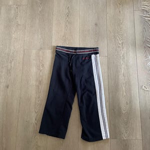 May include: Navy blue sweatpants featuring a white side stripe with thin black lines, a drawstring waistband with red and white horizontal stripes, and the red embroidered text "BCBG" on the upper left side near the waistband.