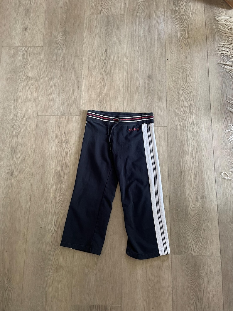 May include: Navy blue sweatpants featuring a white side stripe with thin black lines, a drawstring waistband with red and white horizontal stripes, and the red embroidered text "BCBG" on the upper left side near the waistband.