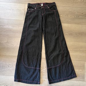 May include: Dark charcoal gray wide-leg denim jeans featuring prominent pink contrast stitching along the seams, pockets, and hem. These pants have a classic button and zipper fly closure, belt loops, and large curved front pockets.