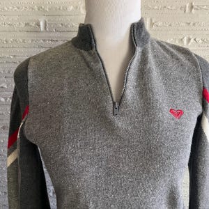 May include: Gray knit sweater with a mock neck and a quarter-zip closure. The sweater features a small red heart logo on the chest and red and white stripes on the sleeves. The sweater is displayed on a mannequin.