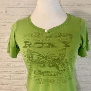 May include: A light green t-shirt with a scoop neck and short sleeves. The shirt features a butterfly graphic and the text "The Original ROXY Boardriding Company". A single white button is at the neckline.