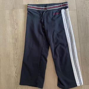 May include: Black cropped sweatpants featuring a white side stripe with thin black lines. The waistband has a drawstring and red and white horizontal stripes. The text BOBG is embroidered in red on the upper left side of the pants.