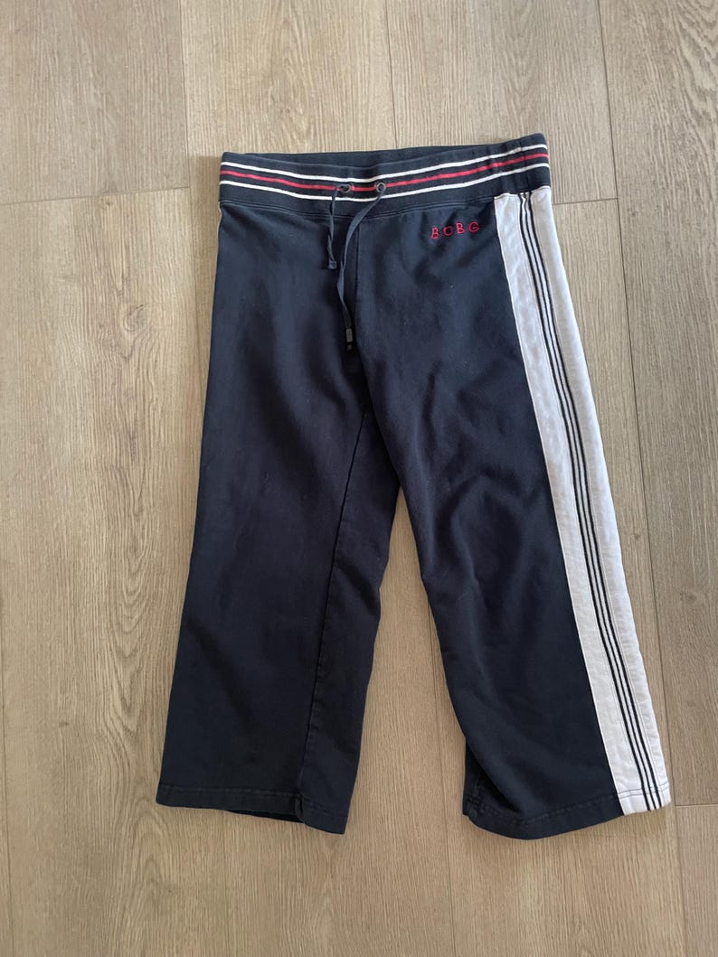 May include: Black cropped sweatpants featuring a white side stripe with thin black lines. The waistband has a drawstring and red and white horizontal stripes. The text BOBG is embroidered in red on the upper left side of the pants.