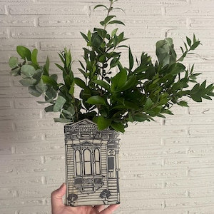 May include: A flat, rectangular ceramic vase featuring a black line drawing of a Victorian-style house facade with a bay window and front door. The vase is filled with a lush arrangement of green eucalyptus and ruscus leaves, creating a full display.