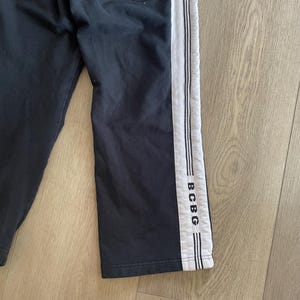 May include: Black sweatpants featuring a wide white vertical stripe down the side of the leg. The white stripe is accented with thin black piping and includes the text BCBG printed in black letters near the bottom hem of the garment.