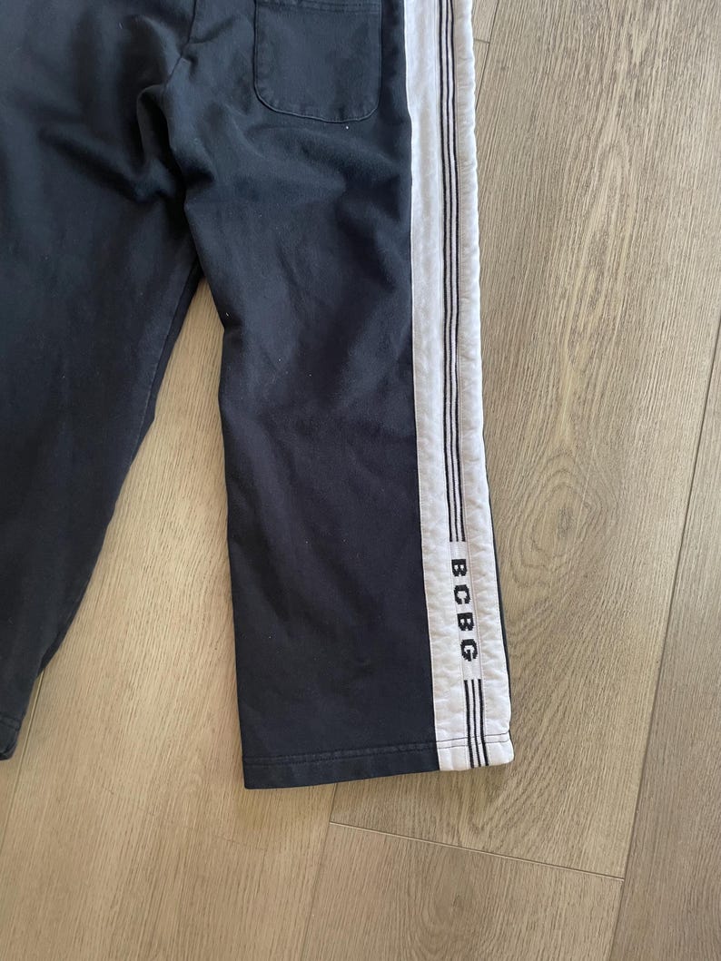 May include: Black sweatpants featuring a wide white vertical stripe down the side of the leg. The white stripe is accented with thin black piping and includes the text BCBG printed in black letters near the bottom hem of the garment.