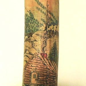 Op de afbeelding: Een kleurrijke tatoeage op de arm van een persoon toont een berglandschap met een klein huisje op de voorgrond. Het huisje heeft een rood dak en een schoorsteen waar rook uit komt. De bergen zijn groen en bruin, en er zijn bomen op de achtergrond.