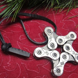 May include: A silver bicycle chain necklace with a star-shaped design. The chain links are connected to form a star shape. The necklace is on a red fabric background with a black leather cord.