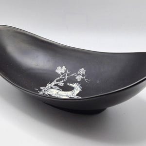 May include: A matte black, boat-shaped bowl with a curved, elongated silhouette. The interior features a white line-art illustration of a deer resting beneath a flowering tree branch, creating a stark contrast against the dark surface.
