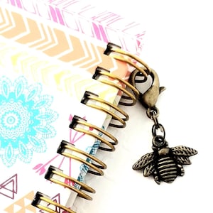 Può includere: Un quaderno a spirale colorato con un motivo geometrico sulla copertina. Un ciondolo a forma di ape in metallo dorato è attaccato al quaderno.