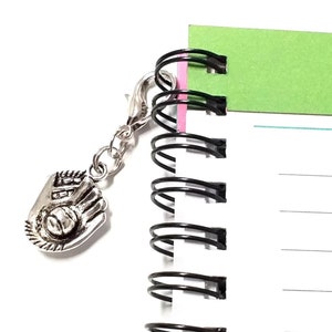 Op de afbeelding: Een zilveren honkbalhandschoenbedel bevestigd aan een zwart spiraalgebonden notitieboek met een groene tab die "MEI" zegt.