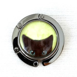May include: A circular, gunmetal-toned metal bag hanger featuring a central glass cabochon insert. The insert displays a graphic of a dark brown cat face with bright blue eyes set against a pale, glowing yellow circular background.