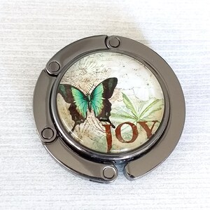 Op de afbeelding: Een zilverkleurige tas haak met een ronde, decoratieve bovenkant met een vlinder met teal en bruine vleugels. Het woord "JOY" is in bruin gedrukt. De achtergrond heeft een vintage kaart ontwerp.