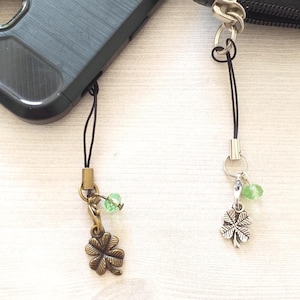 May include: Two phone charms with four-leaf clover pendants. One charm is bronze-toned, the other is silver-toned. Both have green bead accents and black cords. A black phone case and a black wallet are in the background.