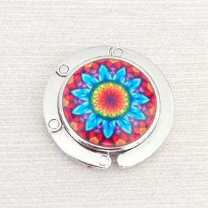 May include: A circular silver-toned metal bag hanger featuring a vibrant, multi-colored mandala design at its center. The pattern displays a bright blue, yellow, orange, and red floral-like geometric arrangement with a glossy, smooth finish.