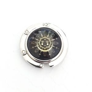 May include: A circular silver-toned metal bag hanger featuring a central glass cabochon. The insert displays a black background with a gold-colored sun face illustration surrounded by radiating starburst patterns and small stars.