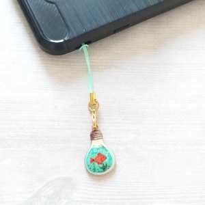 May include: A lightbulb charm with a gold chain and a green string. The lightbulb is filled with a blue liquid and a red fish.