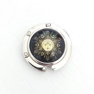 May include: A circular silver-toned metal bag hanger featuring a central glass inlay. The inlay displays a gold-colored sun face graphic surrounded by black rays, stars, and crescent moon symbols, all set within a polished, hinged metallic frame.