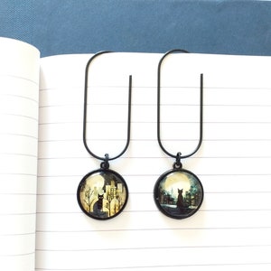 Puede incluir: Dos clips de marcapáginas de metal negro con abalorios redondos de vidrio. Los abalorios presentan una silueta de gato negro frente a un paisaje urbano con luna llena.