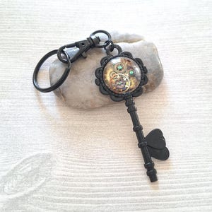 Peut inclure: Porte-clés noir de style steampunk. La clé présente un pendentif circulaire décoratif avec des engrenages et deux pierres vertes. La poignée de la clé a un motif en forme de cœur. Le porte-clés comprend un fermoir et un porte-clés.