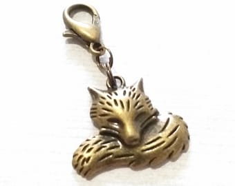 May include: A bronze-toned metal charm shaped like a curled-up fox with etched fur details. The fox charm is attached to a small lobster claw clasp by a circular jump ring, creating a functional accessory piece with a metallic finish.