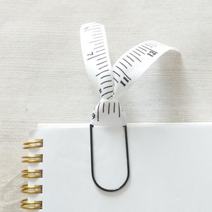 May include: A white measuring tape bookmark with black markings is attached to a black metal binder clip. The bookmark is clipped to a white notebook with gold spiral binding.