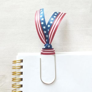 May include: A white notebook with a gold spiral binding and a silver paper clip. The paper clip is decorated with a red, white, and blue ribbon with stars.