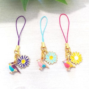 May include: Three colorful phone charms featuring a small bird and a daisy flower. The charms are attached to purple, blue, and pink cords.