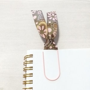 May include: A white notebook with a gold spiral binding and a pink paper clip. A brown ribbon with pink flowers is attached to the paper clip.