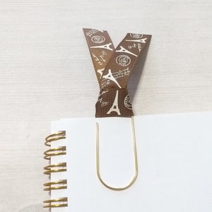 May include: A gold-colored paper clip holds a white spiral notebook open. A brown ribbon with a repeating Eiffel Tower pattern is attached to the paper clip, creating a bookmark. The ribbon is folded over the top of the notebook.