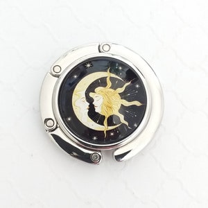May include: A circular silver-toned metal bag hanger featuring a central inlay with a black background. The design depicts a yellow crescent moon and a golden sun with human-like faces facing each other, surrounded by small white stars.