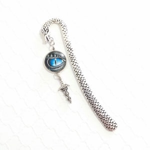 Op de afbeelding: Een zilverkleurige boekenlegger met een drakenhuidpatroon. Het heeft een drakenoog-hanger met een blauwe iris en een kleine zwaardbedel. De boekenlegger heeft een gebogen bovenkant met een drakenkop.