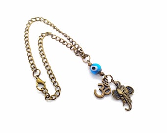 May include: A bronze-toned metal chain featuring a blue and white glass evil eye bead, a bronze Om symbol charm, and a bronze elephant head charm. The chain includes a lobster claw clasp, creating a decorative accessory with multiple charms.