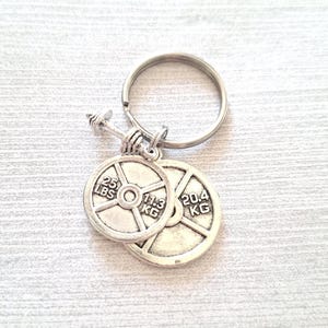 May include: Silver-tone keyring with a dumbbell charm and two weight plate charms. One plate is marked "25 LBS" and "11.3 KG", the other is marked "20.4 KG". A fitness-themed accessory.