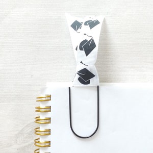 May include: A white notebook with a gold spiral binding and a black metal bookmark. The bookmark is decorated with a white ribbon with black graduation cap designs.