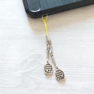 May include: A silver tennis racket phone charm with a yellow string attached to a black phone case.