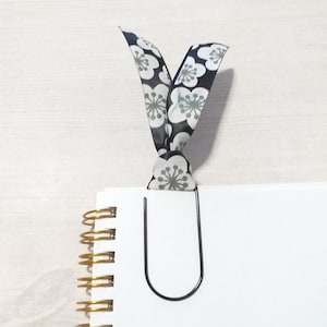 May include: A black metal paper clip with a black and white floral ribbon attached to it. The ribbon is tied in a bow and the paper clip is holding a blank white page.