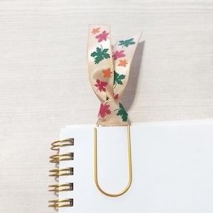 May include: A gold-colored paper clip with a cream-colored ribbon bookmark. The ribbon is printed with colorful autumn leaves in shades of green, orange, and pink. The bookmark is attached to a white notebook.