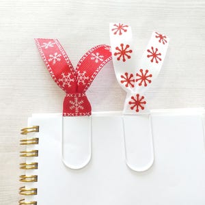 May include: Two white metal paper clips with red and white ribbon bookmarks. The ribbons have a snowflake pattern.