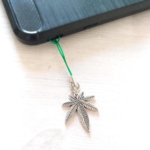 May include: A silver charm in the shape of a marijuana leaf hangs from a green string attached to a black smartphone.