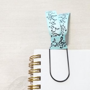 May include: A white notebook with a gold spiral binding and a black metal bookmark. The bookmark is attached to a light blue ribbon with black musical notes printed on it.