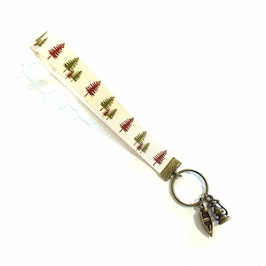 May include: A white ribbon keychain with a pattern of green and red trees. The keychain has a metal ring with a small metal boat and lantern charm.