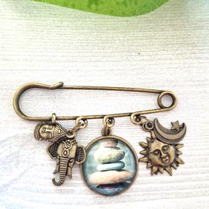 Puede incluir: Un broche de alfiler de seguridad en tono bronce con tres colgantes. Los colgantes incluyen un elefante, un colgante redondo con piedras apiladas y un diseño de sol y luna. El alfiler está sobre una superficie de madera blanca.
