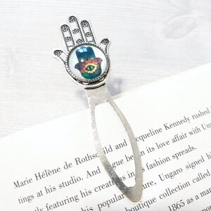May include: Silver-toned metal bookmark featuring a Hamsa hand design with an eye symbol. The Hamsa hand has a detailed, openwork design. The eye symbol has blue, green, and yellow colors. The bookmark is placed on a book.