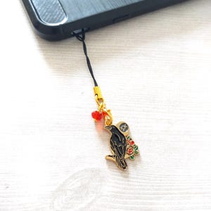 May include: A black and gold phone charm depicting a raven on a branch with red and green floral details. The charm is attached to a black cord and a gold-coloured metal clasp.